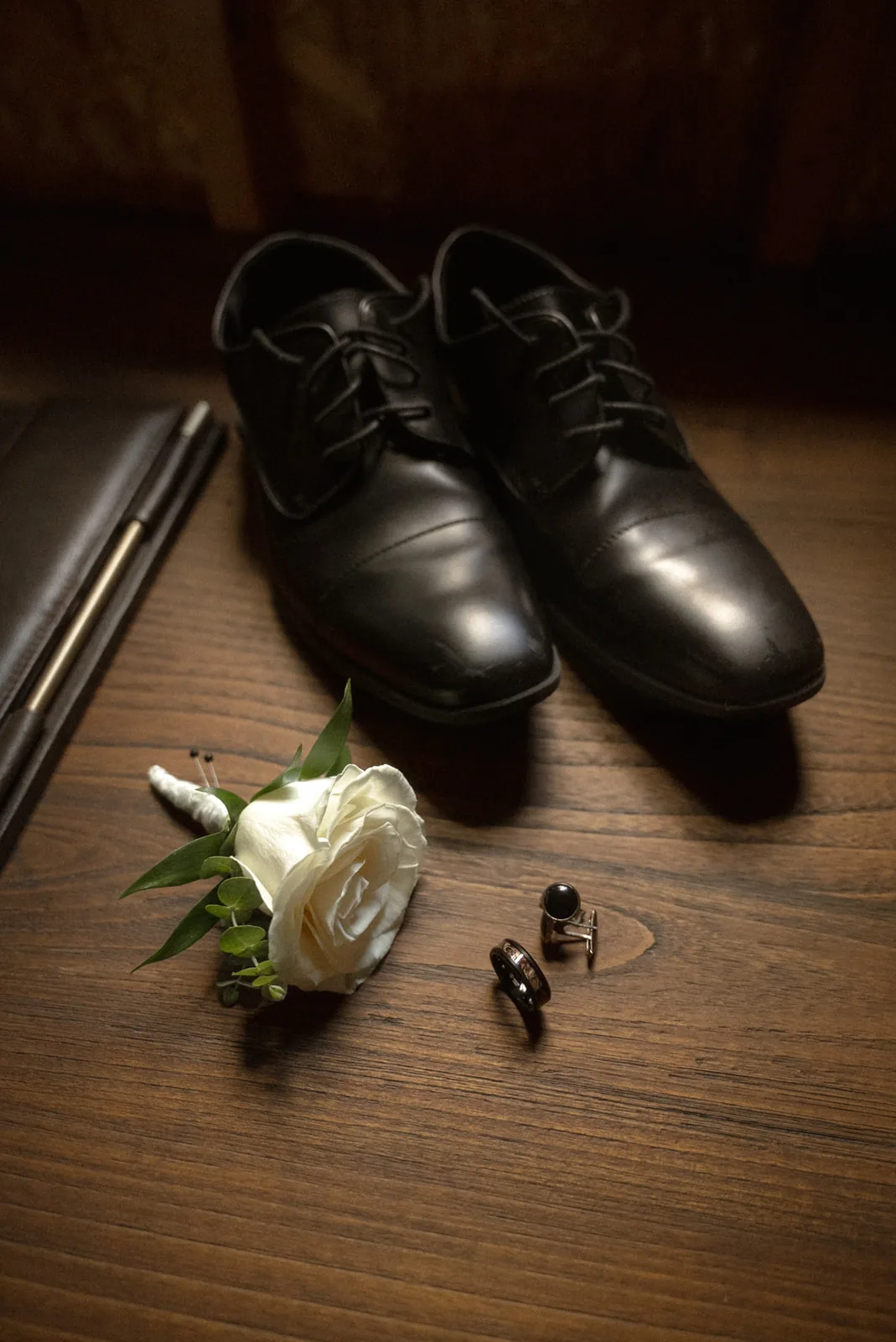 detail flatlay of groom shoes, boutonniere, rings, pen on wood table