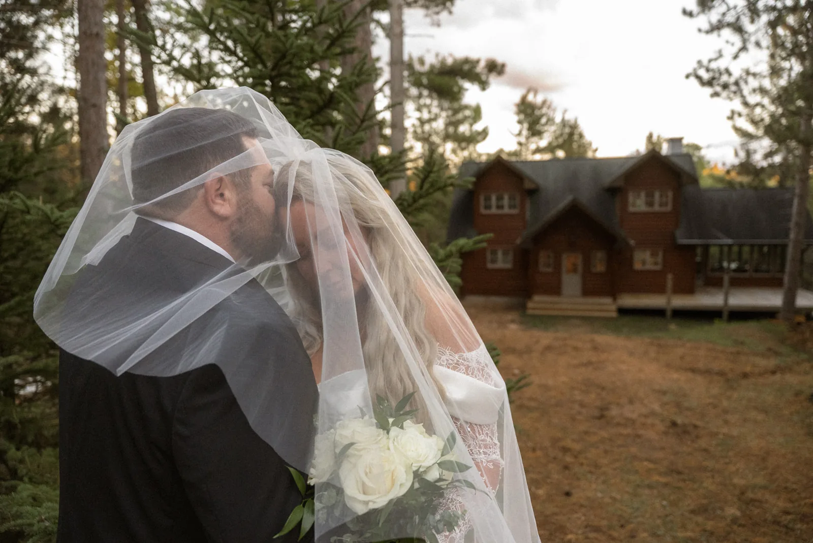 groom kissing bride under veil, cabin in background