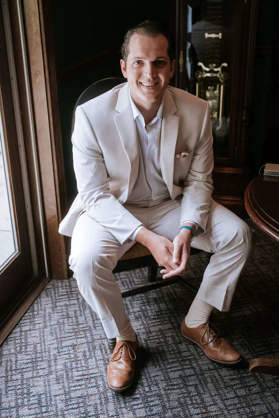 groom portrait seated in tan suit, sunroom window light