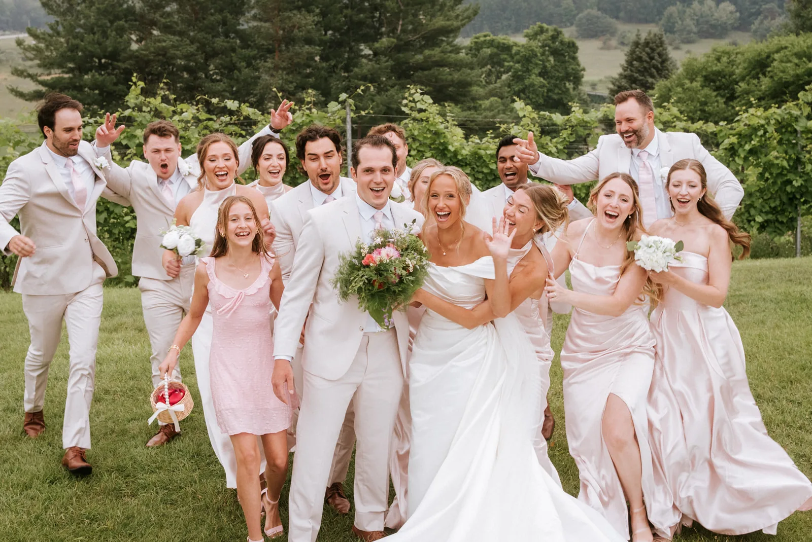 wedding party candid walking toward camera laughing, bouquets raised, vineyard
