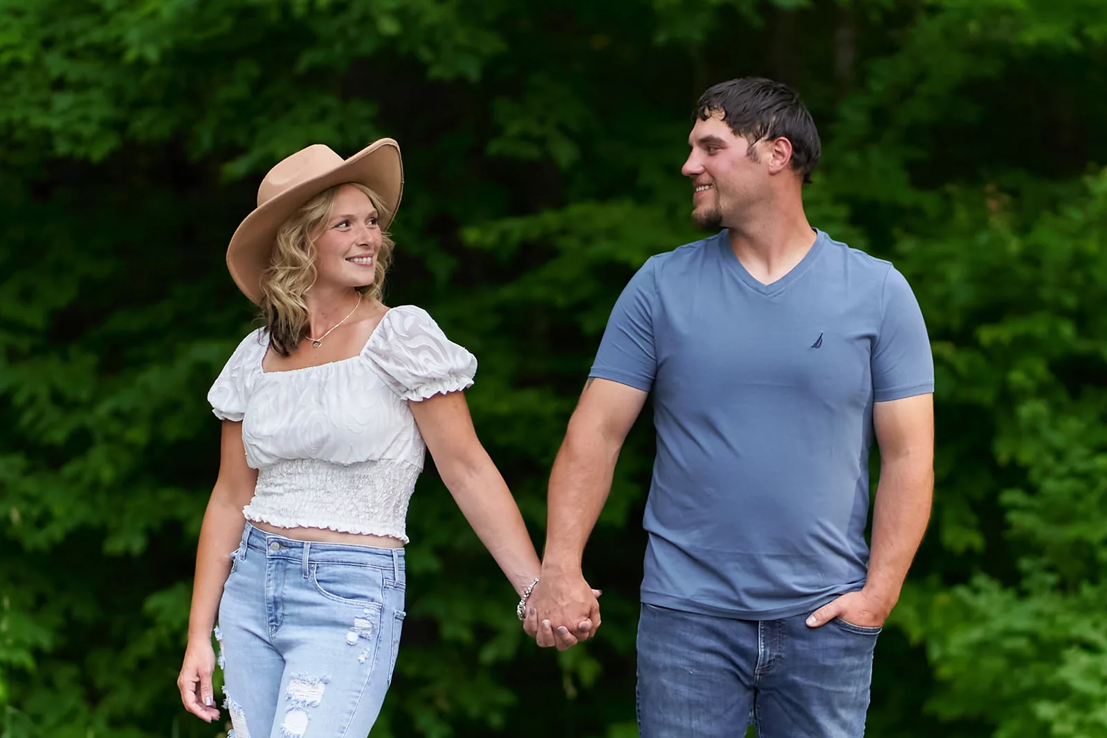 couple walking hand in hand through green forest, cowboy hat