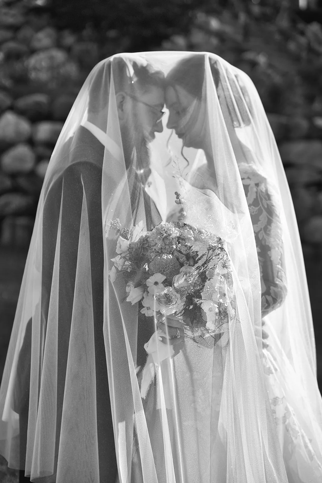 bw bride standing under flowing veil with bouquet, stone wall backdrop