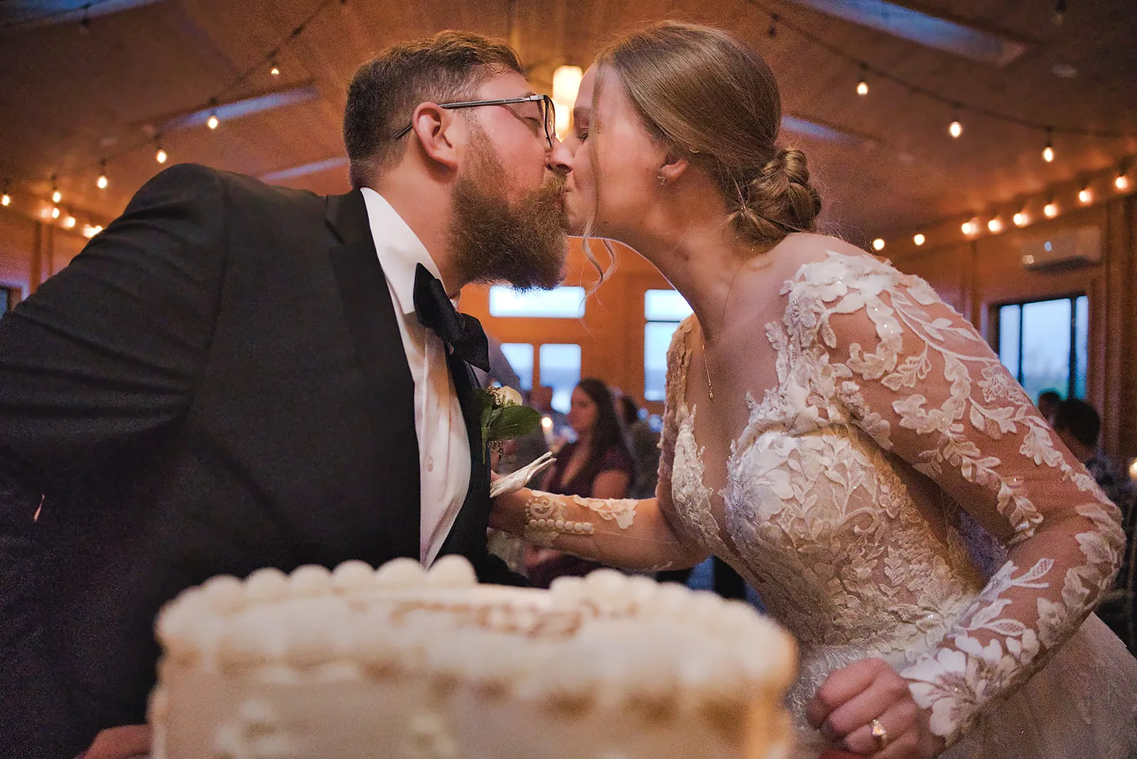 bride and groom kissing while cutting cake, close-up candid, string lights