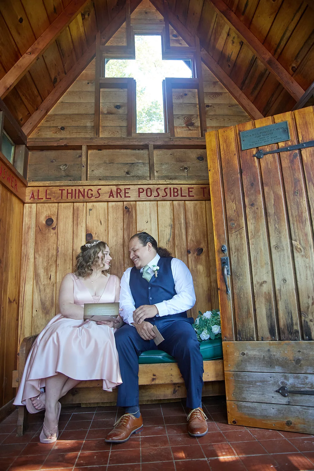 bride and groom seated in small wooden chapel with cross window