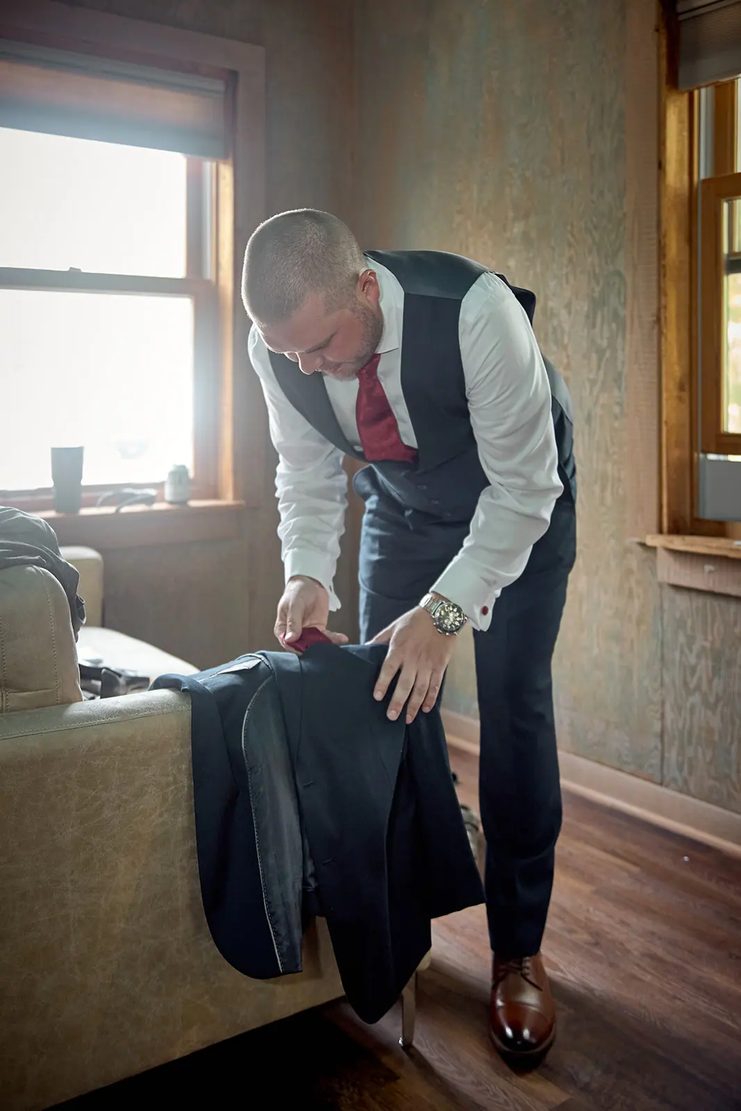 Groom folding jacket sunlit window — Taylor & Mark's wedding at The Manor at Fire Tower Hill, Lewiston, MI
